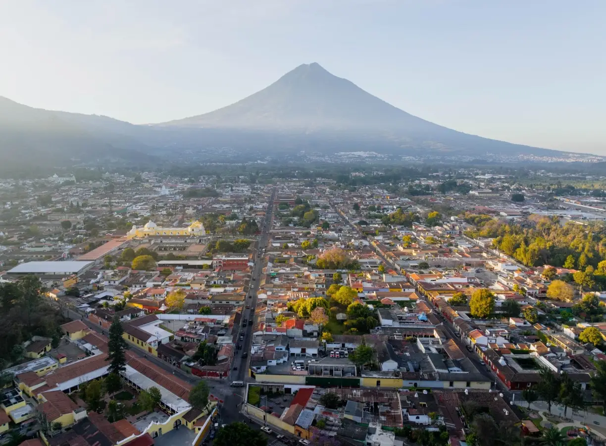 Aerial drone photograph of Antigua Guatemala city grid and colonial buildings with Volcan de Agua peak in the background under a hazy sky.