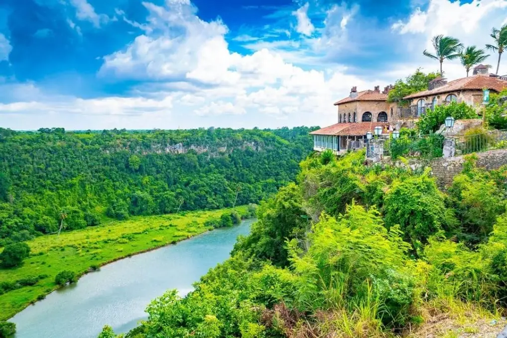 Mediterranean-style stone buildings of Altos de Chavón perched on a lush green cliffside above a wide winding river in La Romana, Dominican Republic.