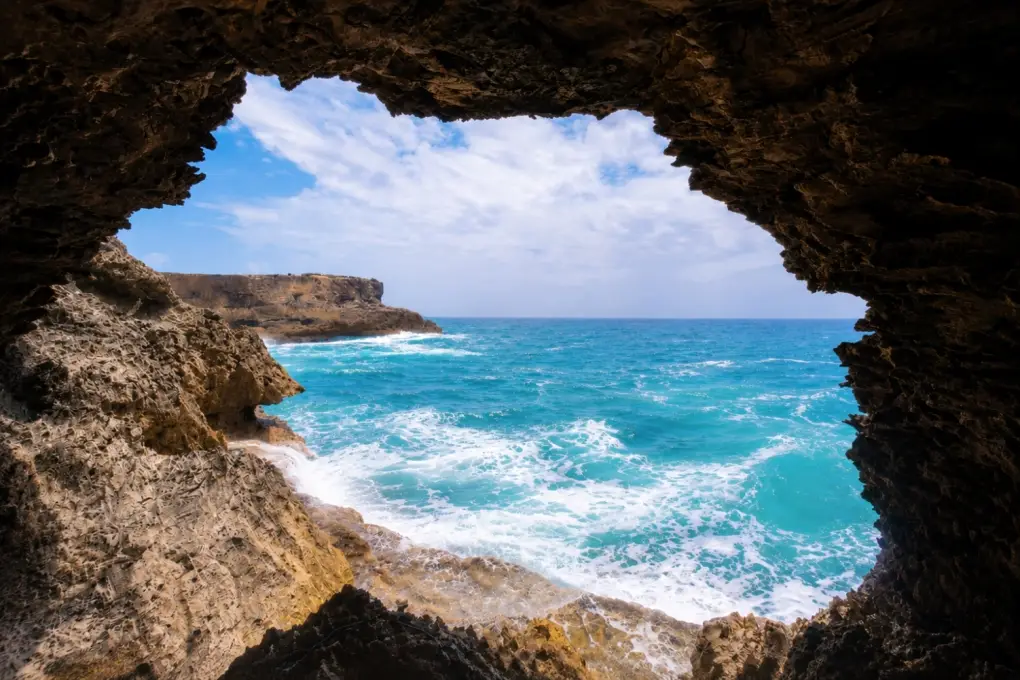 Looking out from inside Animal Flower Cave in Barbados at rough turquoise ocean waves through a natural rock opening under a blue sky.