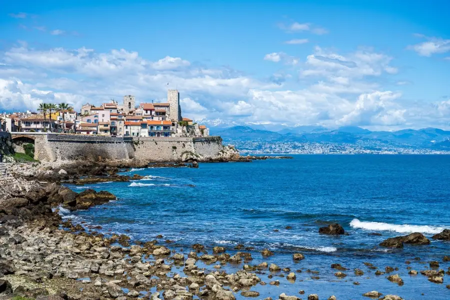 The historic walled seaside town of Antibes on the French Riviera, featuring stone ramparts, historic brick buildings and clear blue Mediterranean waters with snow-capped Alps in the background.