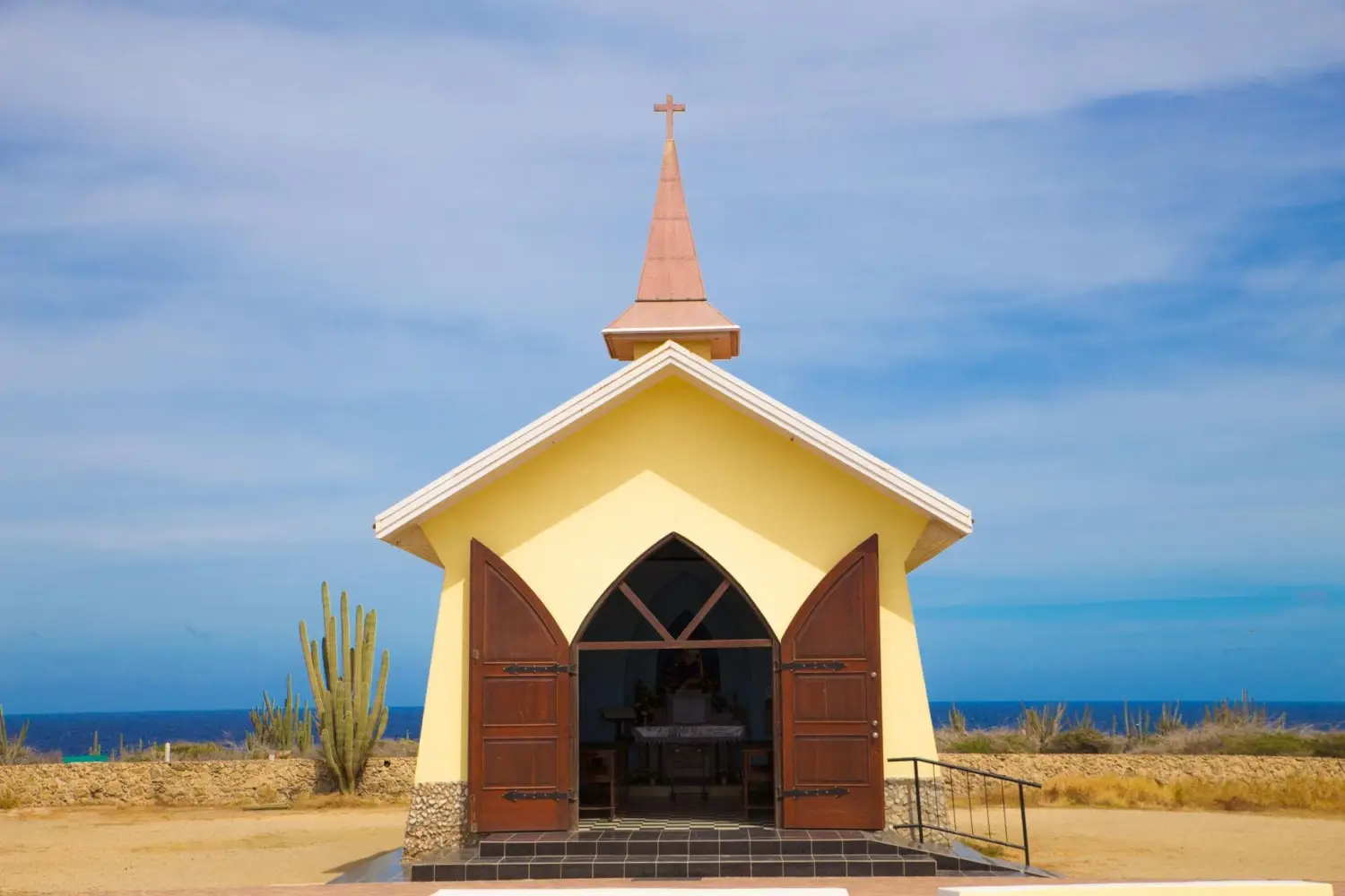 Alto Vista Chapel is a small, bright yellow Roman Catholic chapel with scenic coastal views in Aruba surrounded by cacti under a blue sky.