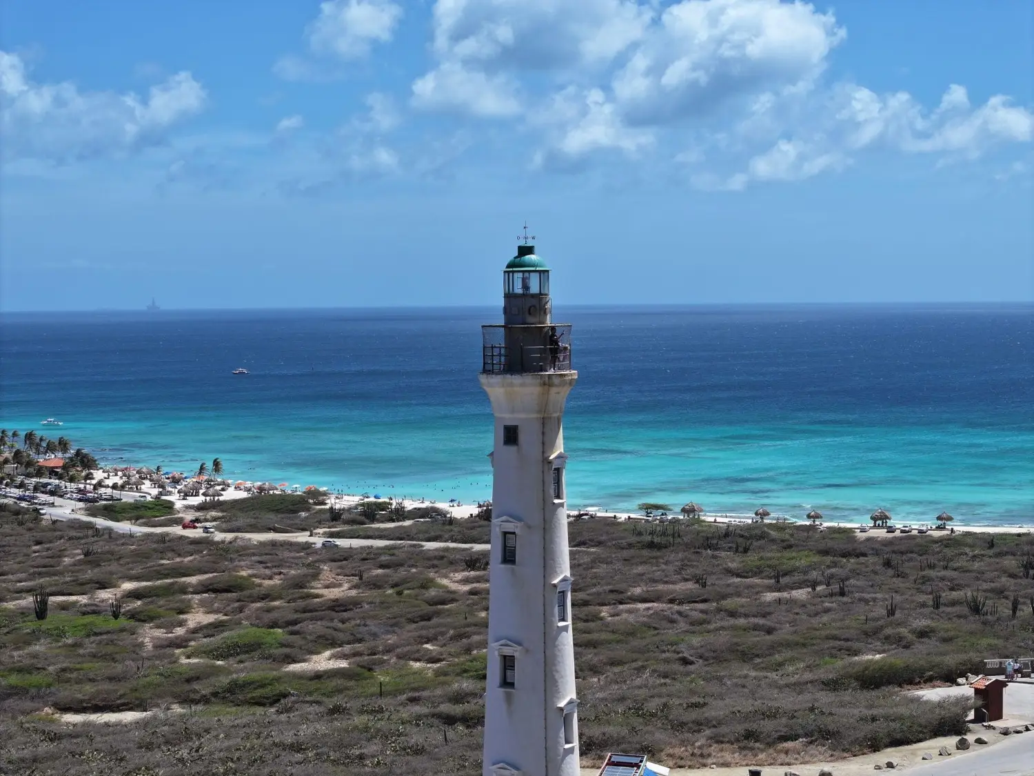 The white stone tower of the historic California Lighthouse near Arashi Beach in Aruba standing tall on a grassy hill overlooking a vast blue ocean under a cloudy sky.