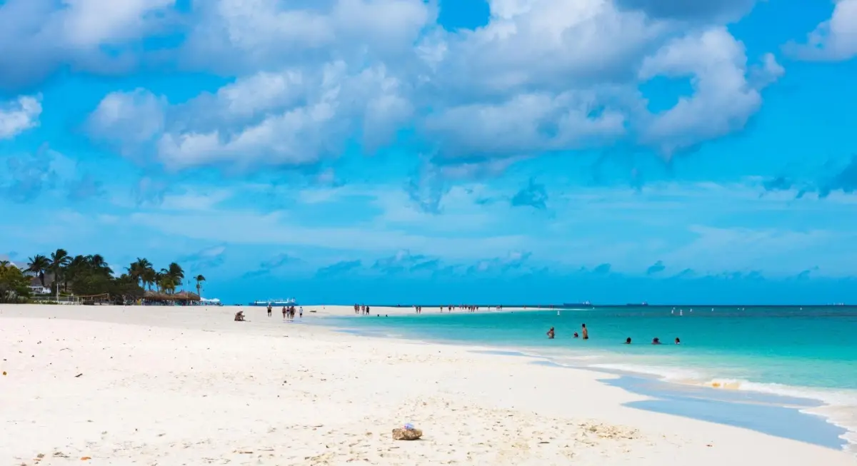 A wide panoramic view of Eagle Beach in Aruba featuring soft white coral sand, vibrant turquoise sea, and people swimming under a partly cloudy blue sky, frequently voted one of the world's best beaches.