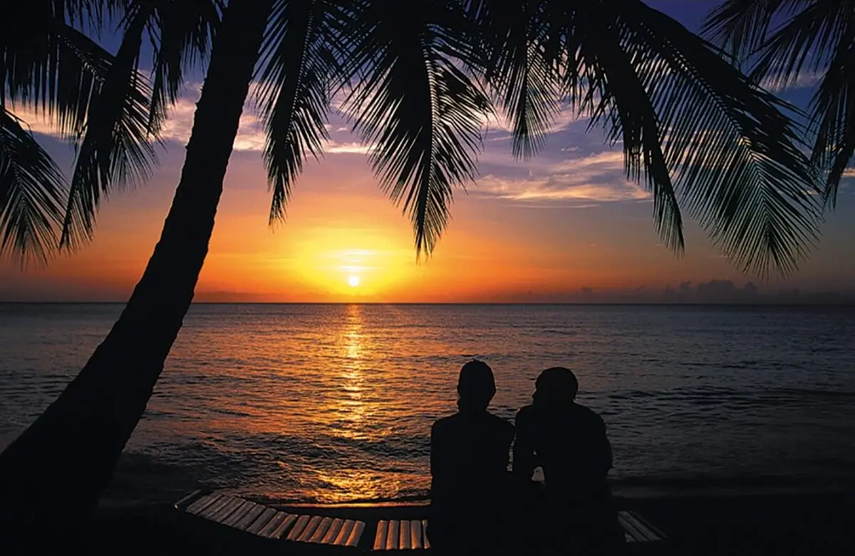 Silhouetted couple watching a golden sunset on a tropical beach in Barbados under palm trees.