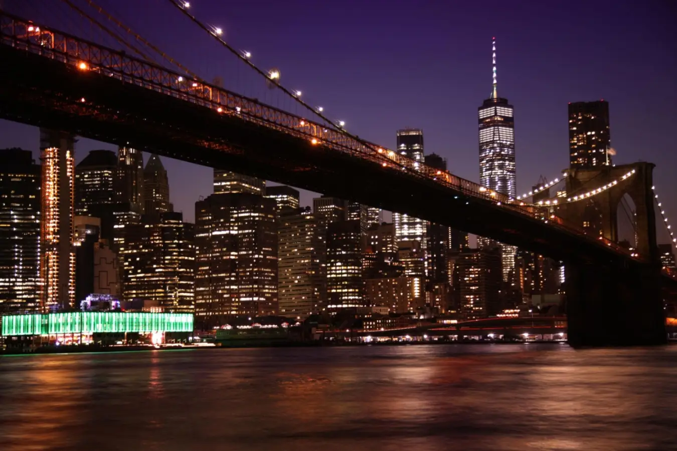 The illuminated Brooklyn Bridge spanning across the East River in New York City showcasing the Lower Manhattan skyline at night.