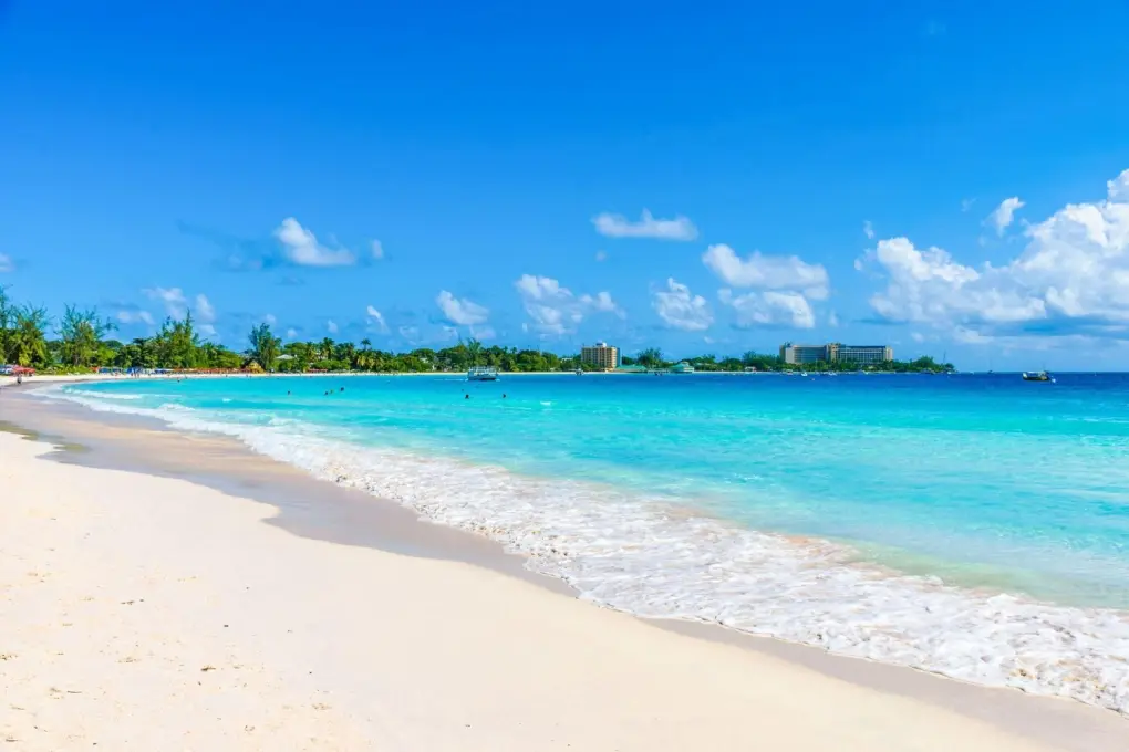 Wide scenic view of the white sand beach and turquoise ocean water at Carlisle Bay in Barbados under a blue sky with light clouds.
