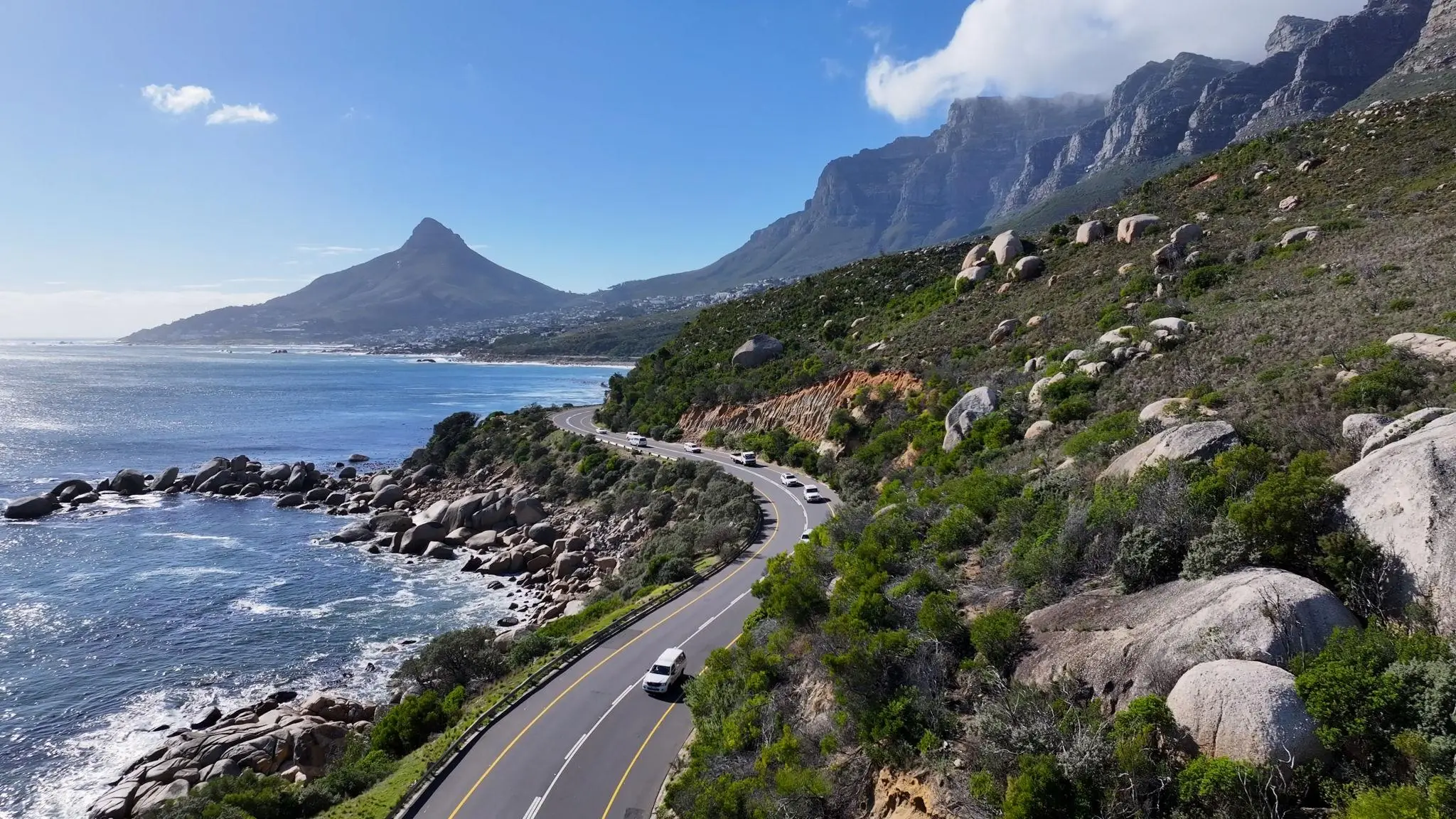 A winding coastal highway in Cape Town, South Africa, with cars driving between the ocean and rocky, green mountain slopes.