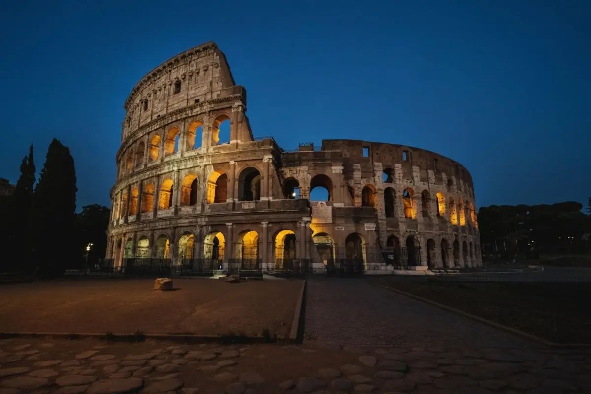 Exterior view of the illuminated ancient Roman Colosseum amphitheater arches against a dark blue evening sky.