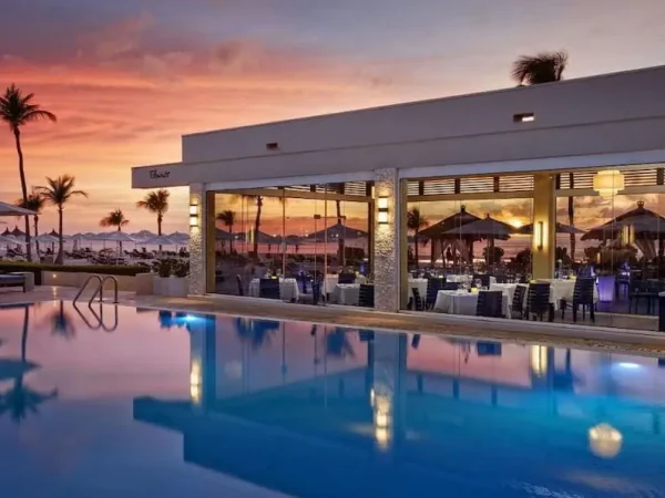 Bucuti & Tara Beach Resort in Aruba showing a luxury infinity pool reflecting a vibrant sunset next to Elements Restaurant's indoor dining area.