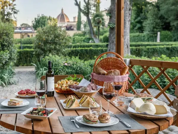 A luxury outdoor picnic set up at the Four Seasons Hotel Firenze in Florence, Italy, featuring a wooden table laden with sandwiches, cheeses, wine, and a wicker basket.