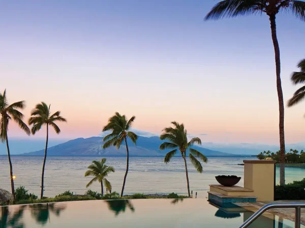 An infinity edge swimming pool reflects towering palm trees at dusk on Maui, Hawaii. In the background, a calm ocean stretches out toward a mountainous island silhouette under a soft pink, purple, and blue gradient sky.