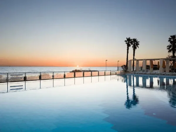 An outdoor infinity pool reflecting palm trees and a clear blue sky at sunset, overlooking a glass railing that separates the pool deck from the open ocean in Malta.