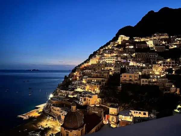 Night view of Positano on the Amalfi Coast in Italy, showing the cliffside village illuminated with warm lights overlooking the dark blue Tyrrhenian Sea under a twilight sky.