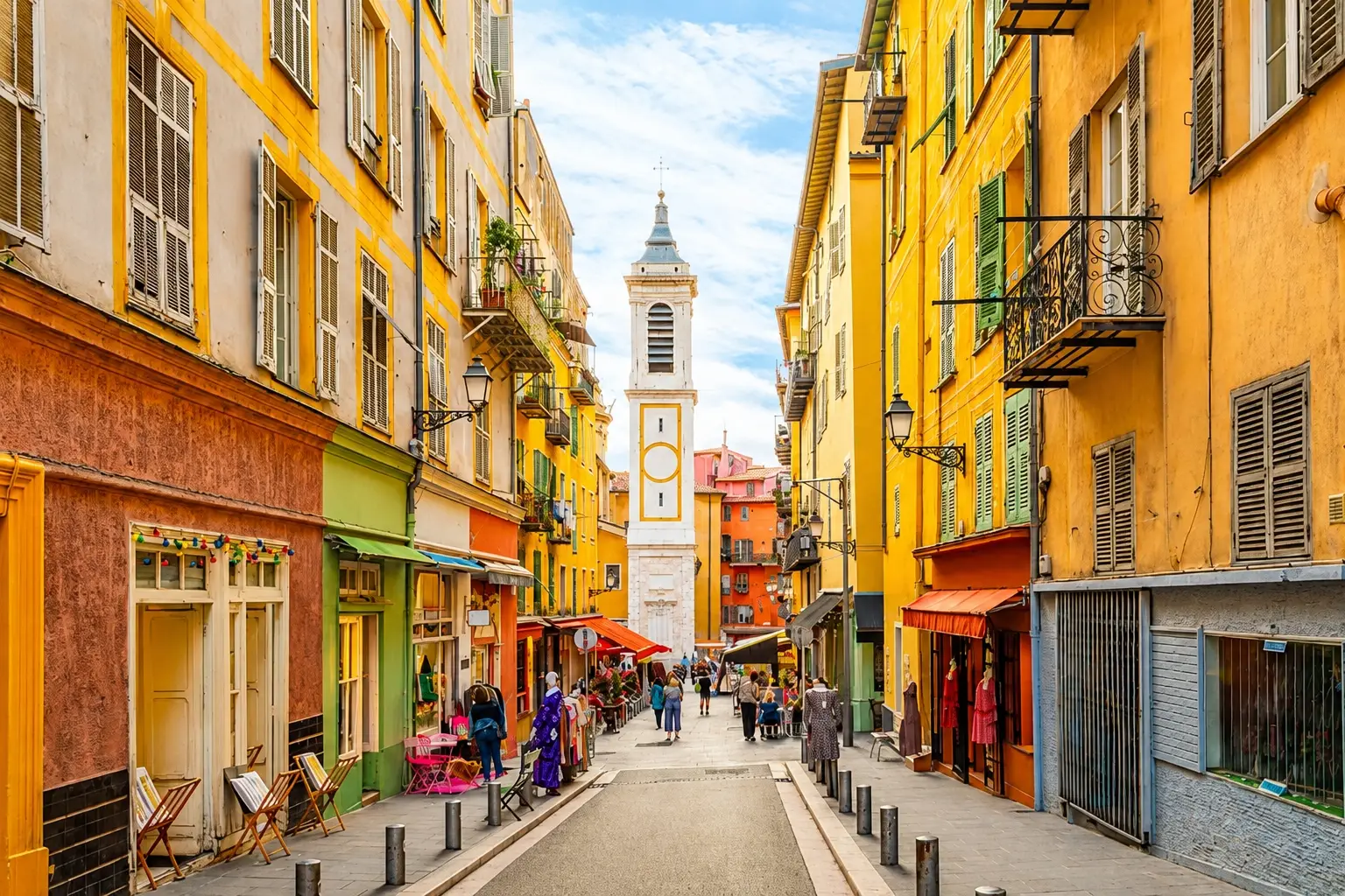 A sunny, pedestrian only street within the Old Town of Nice in France, architecturally characterized by tall, narrow apartment buildings painted in vibrant yellow, orange and red Mediterranean hues.