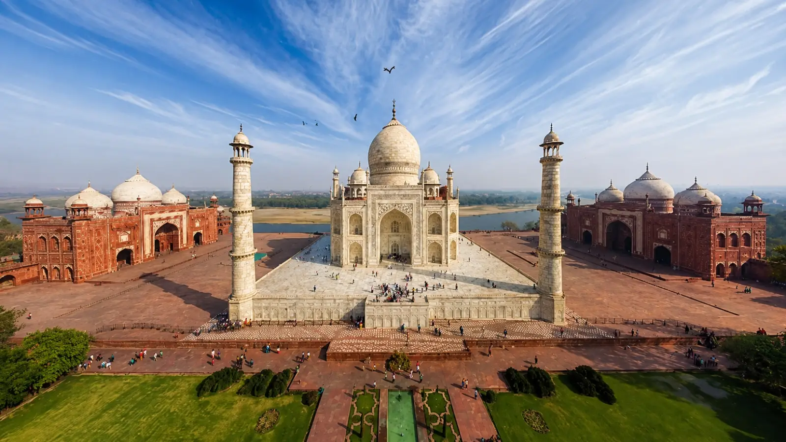 Aerial view of the Taj Mahal complex in Agra India showcasing the main white marble mausoleum two red sandstone buildings on either side four corner minarets and front gardens under a blue sky.