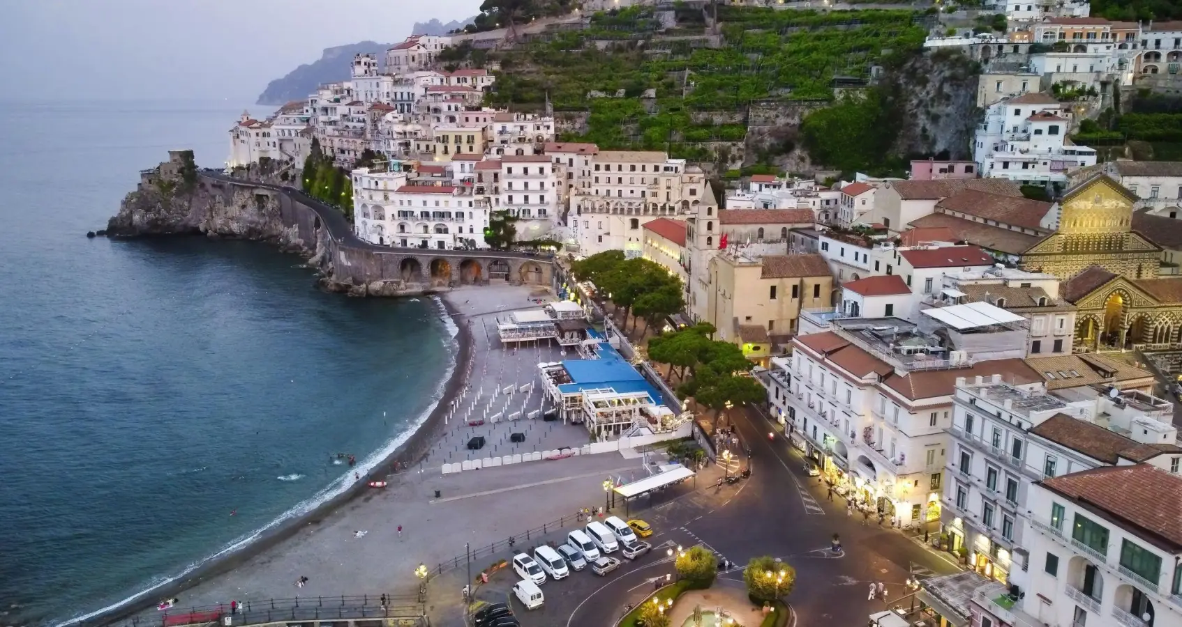 Aerial view of Amalfi town beach and coastline with white buildings on steep cliffs at dusk, Campania, Italy.