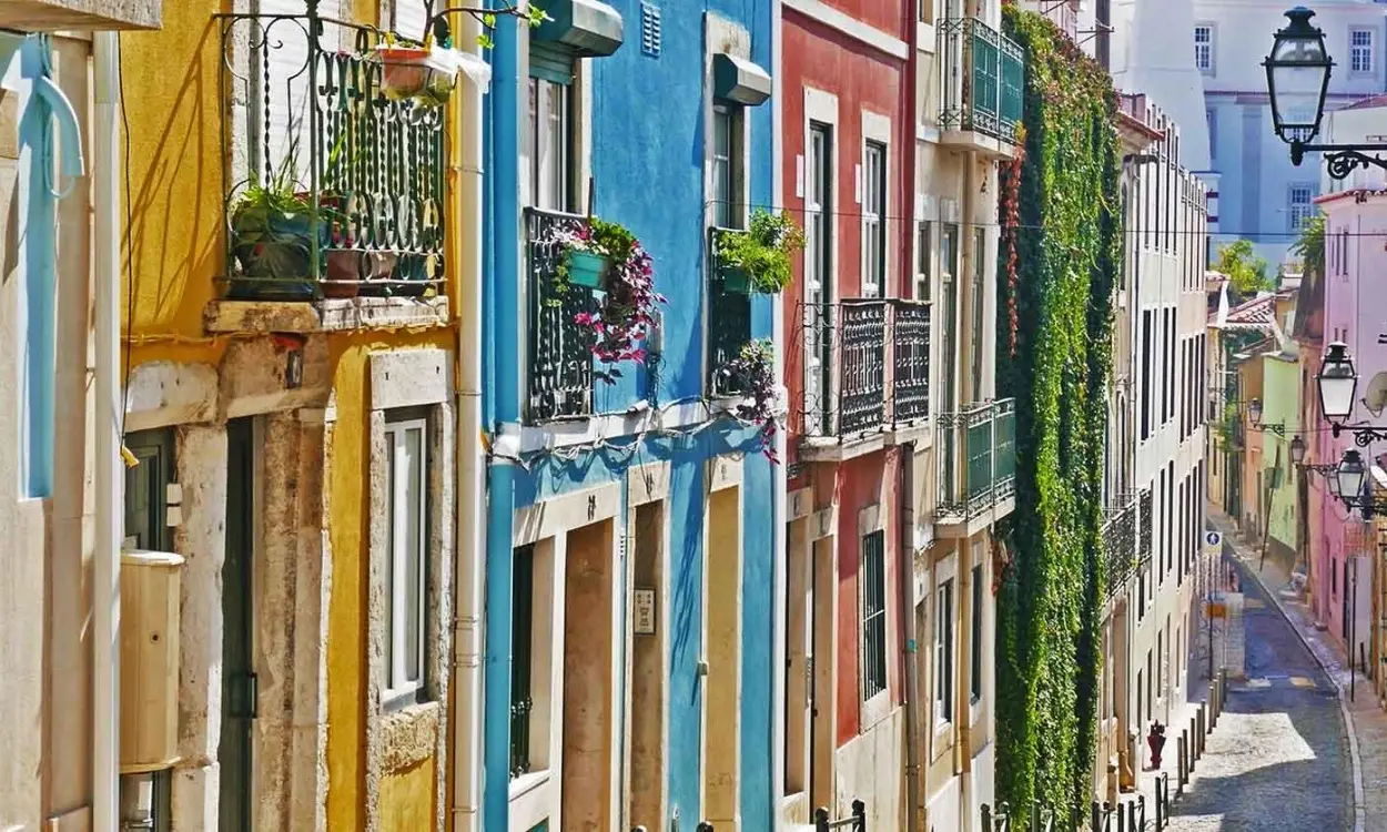 Side view of a narrow sloping street in Lisbon with yellow, blue, and pink historic apartment buildings, wrought-iron balconies with potted flowers, and an ivy-covered wall.
