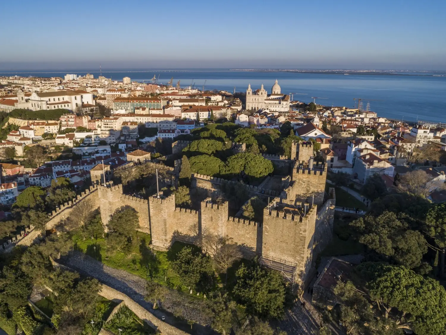 Panoramic view from Castelo de Sao Jorge in Lisbon, Portugal, showing historic rooftops, the Tagus River, and the National Pantheon dome under a clear blue sky.
