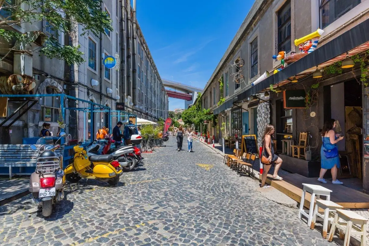 A cobblestone street at LX Factory in the Alcantara neighborhood of Lisbon, Portugal, featuring historic industrial buildings, local shops, and outdoor cafes.