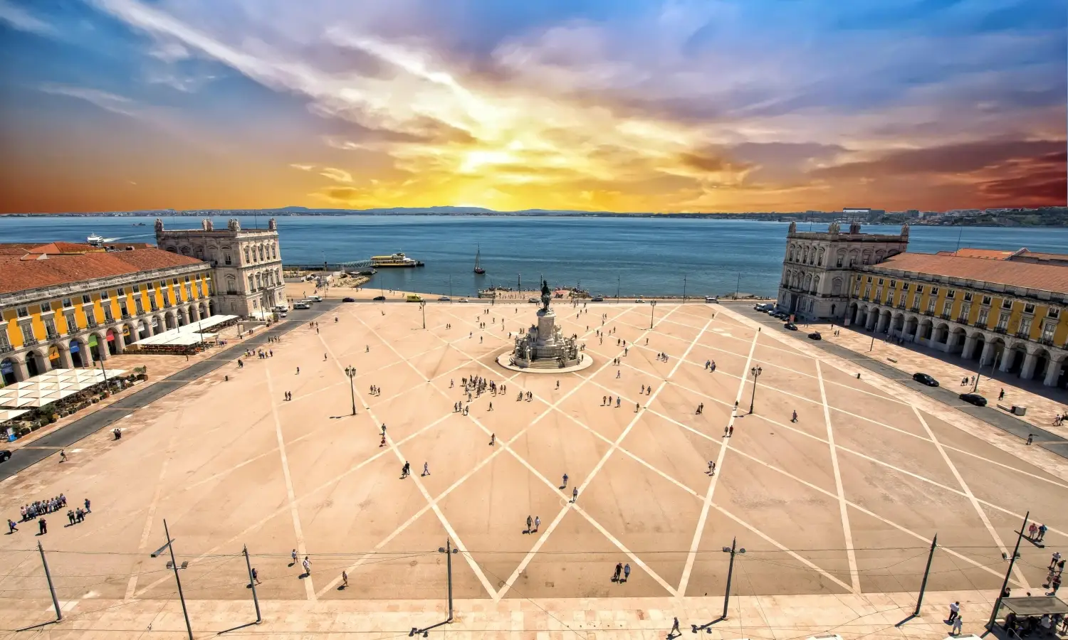 Aerial view of Praca do Comercio plaza in Lisbon, Portugal, featuring yellow arcaded buildings, a statue of Dom Jose I, and the Tagus River at sunset.