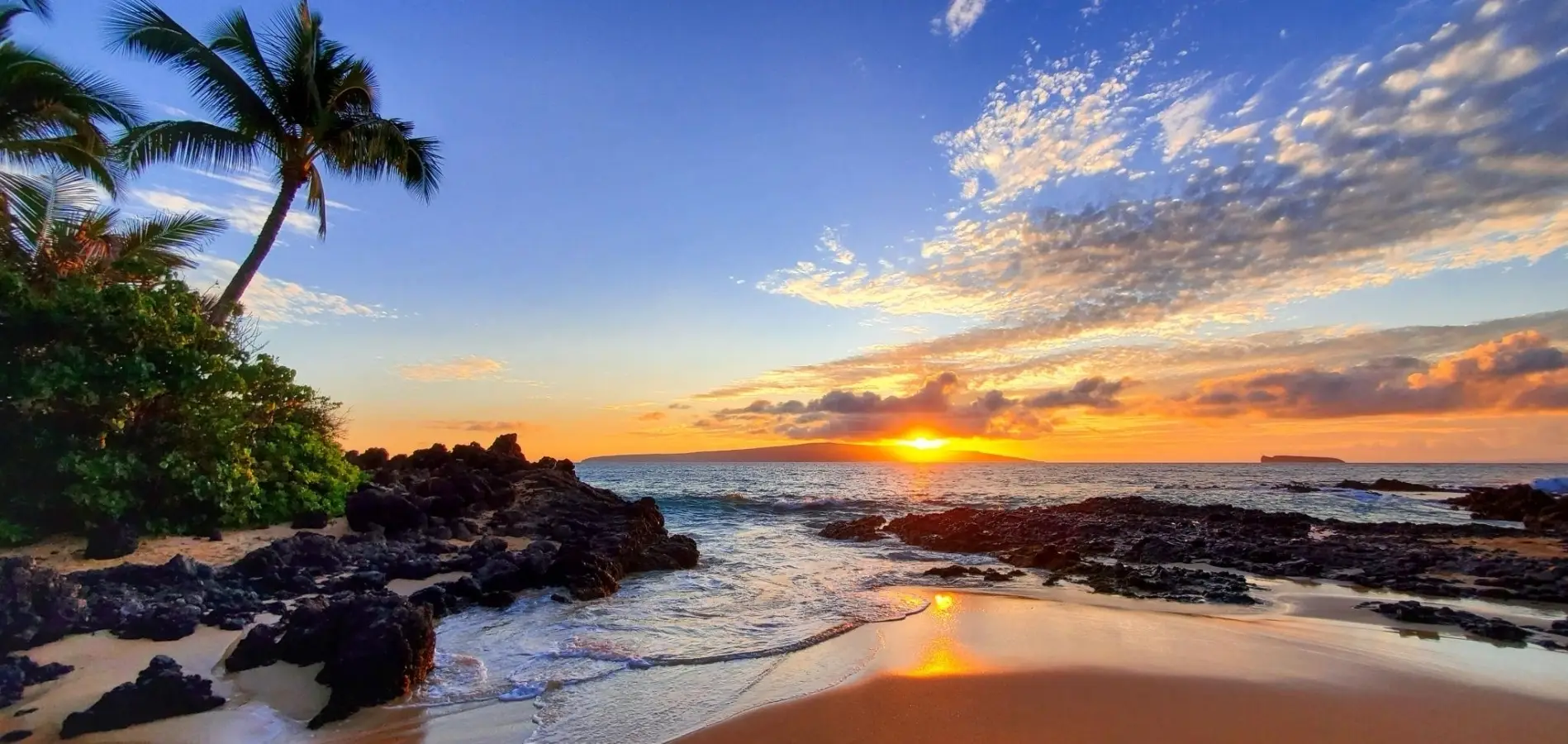Sunset over the ocean at Makena Secret Beach in Maui Hawaii with volcanic rocks and palm trees.