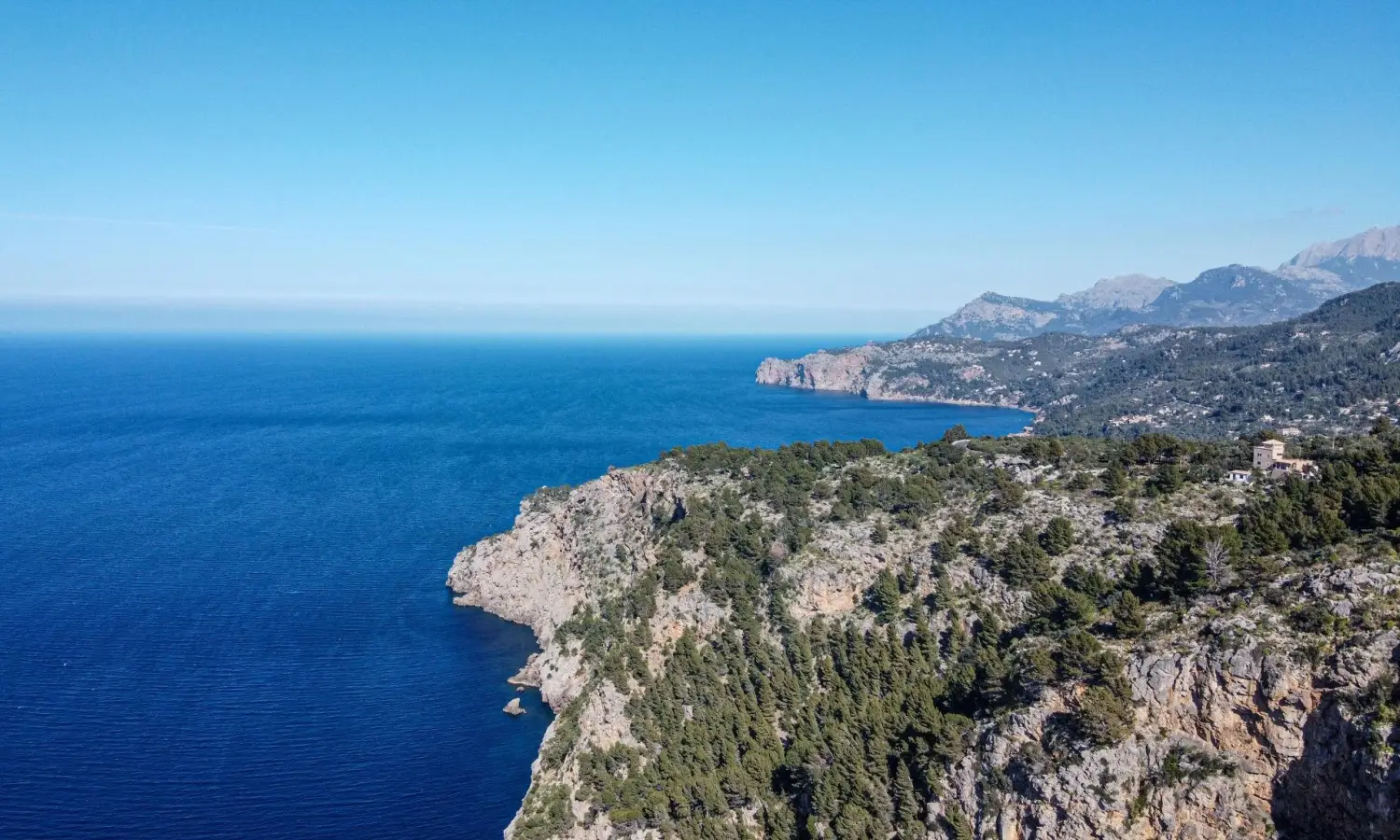 Rugged limestone cliffs and pine-covered hills of the Serra de Tramuntana mountains meeting the blue Mediterranean Sea in Mallorca, Spain.