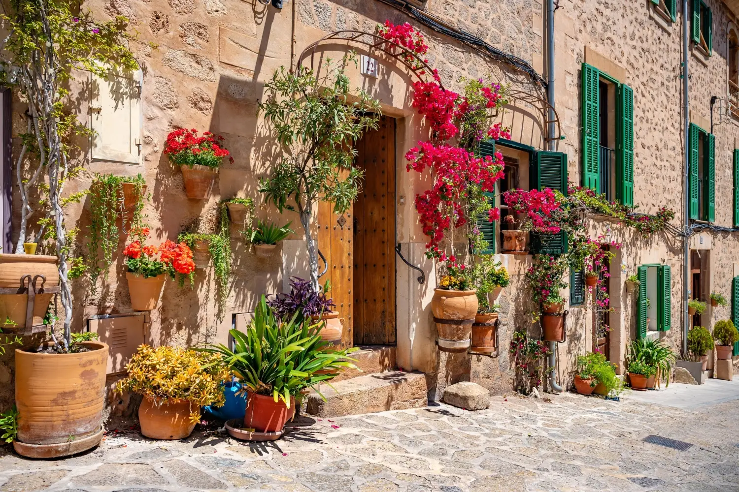Traditional stone building in Valldemossa, Mallorca, decorated with bright pink bougainvillea, terracotta pots, and green shutters.