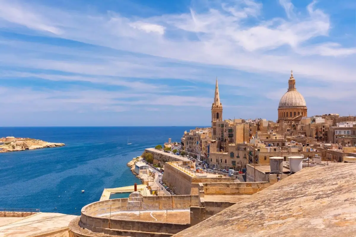 A panoramic view of Valletta, Malta featuring the historic Carmelite Church dome and St Paul's Pro-Cathedral spire against a bright blue Mediterranean Sea and cloudy sky.