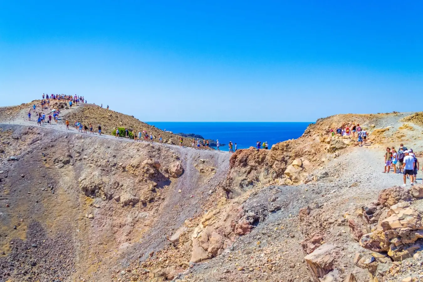 Tourists hiking along the rocky volcanic ridge trail of Nea Kameni island in Santorini Greece under a clear blue sky.