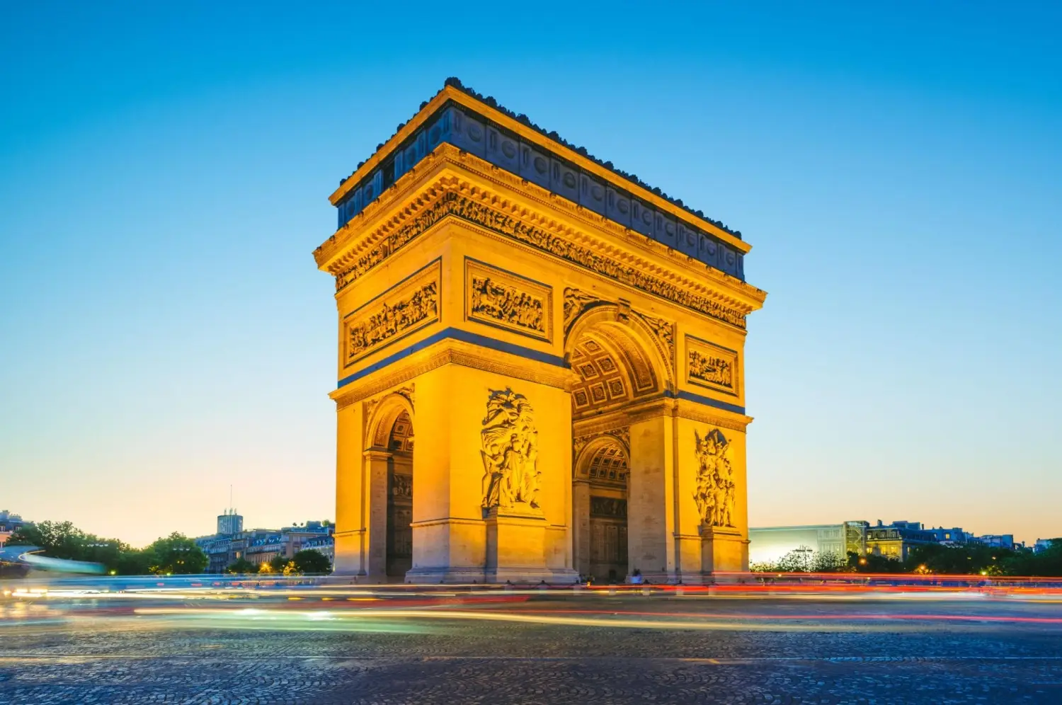 The Arc de Triomphe at the western terminus of the Champs-Elysees in Paris, standing illuminated at twilight at the center of Place Charles de Gaulle.