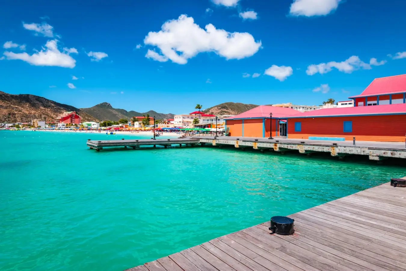 Vibrant wooden dock over turquoise ocean water at Great Bay Beach in Philipsburg, Sint Maarten, featuring colorful tropical buildings under a blue sky.