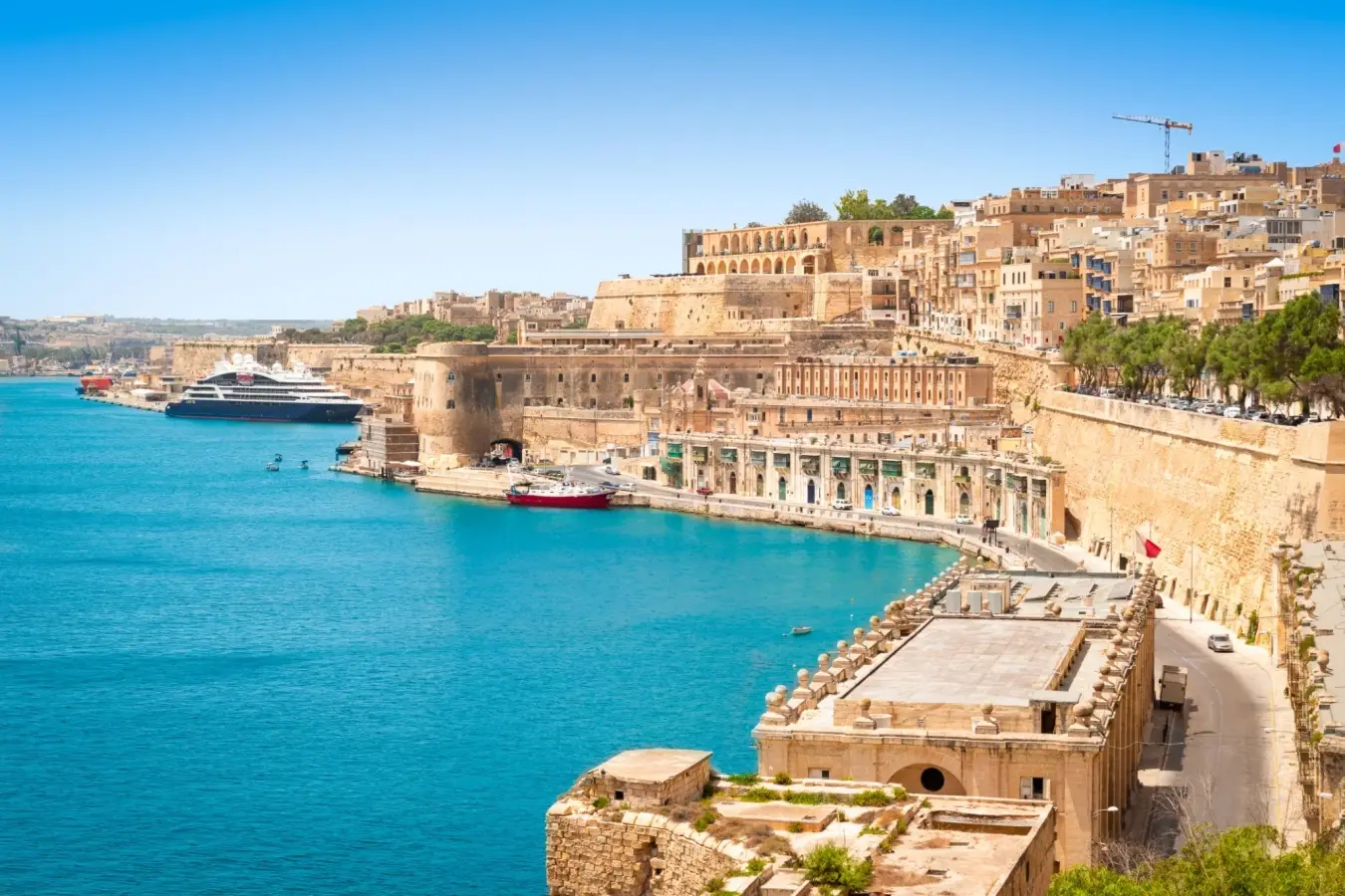 Panoramic view of the Valletta Grand Harbour in Malta, featuring medieval limestone city walls, historic coastal buildings, vibrant blue water, and a cruise ship docked in the background under a clear sky.