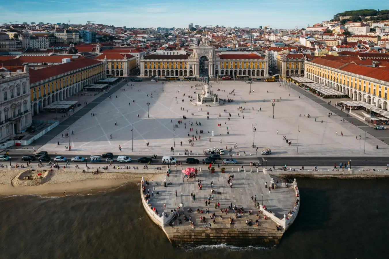Aerial view of Praca do Comercio public square in Lisbon, Portugal, showing the neoclassical architecture, yellow buildings, Rua Augusta Arch, and crowds of pedestrians next to the Tagus River waterfront.