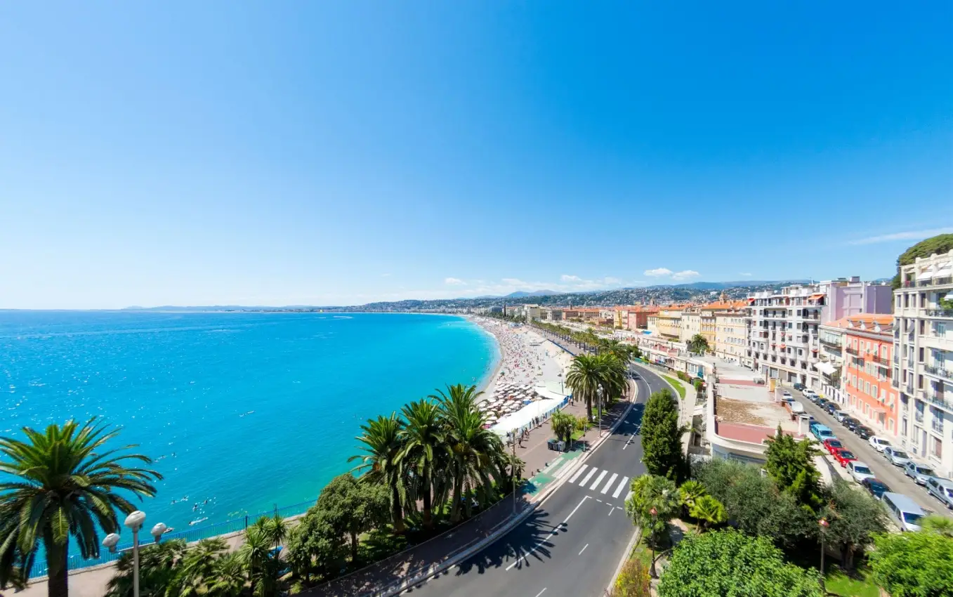 Aerial view of the Promenade des Anglais in Nice France showing blue Mediterranean sea, pebble beach, palm trees, and coastal road.