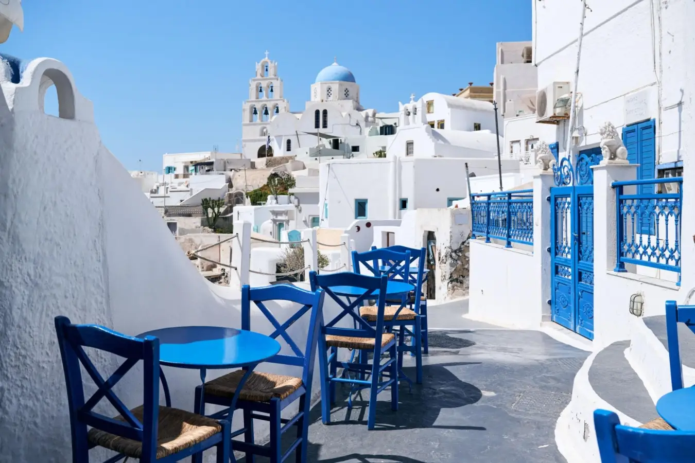 Blue wooden tables and chairs set outside along a narrow, white stone pathway overlooking the iconic white buildings and blue-domed church of Pyrgos village in Santorini, Greece.