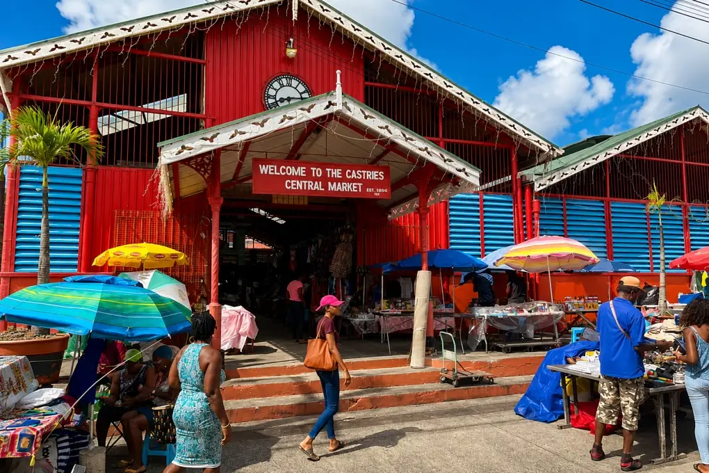 Bustling crowd walking past the bright red and blue Castries Central Market entrance in Saint Lucia under a sunny blue sky.