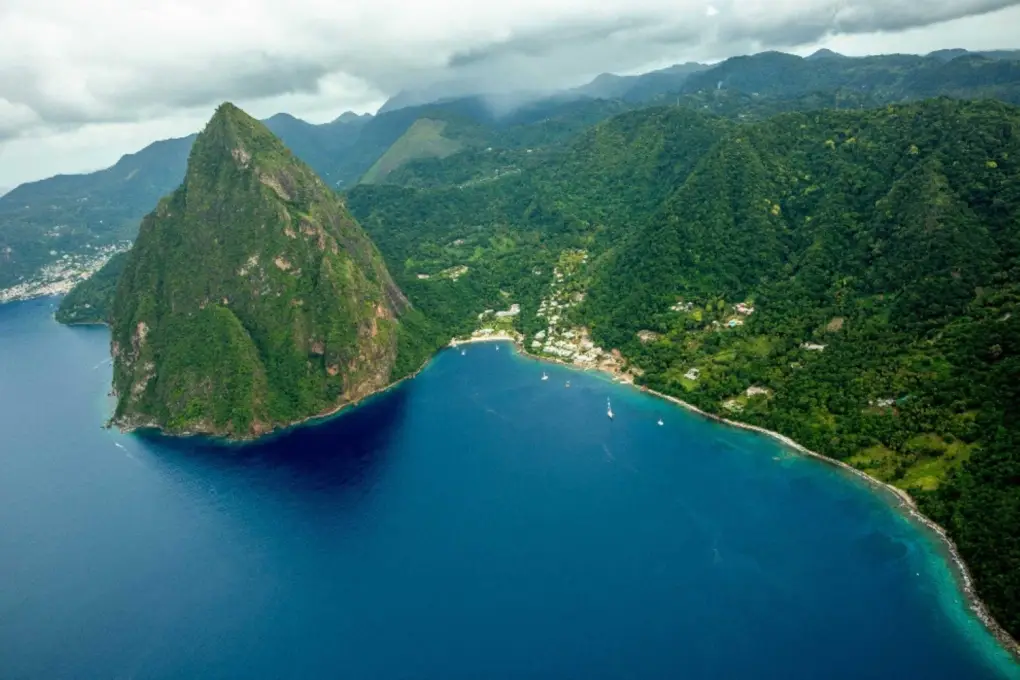 Petit Piton, a lush, green volcanic mountain peak rising sharply from clear blue Caribbean water along the forested coastline of Saint Lucia under a cloudy sky.