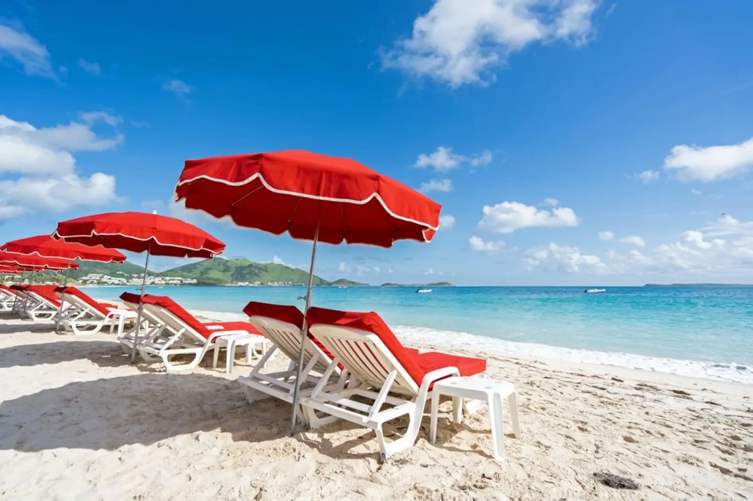 White sand beach with a row of vibrant red umbrellas and white lounge chairs facing turquoise water under a blue sky at Orient Bay Beach in St Martin.