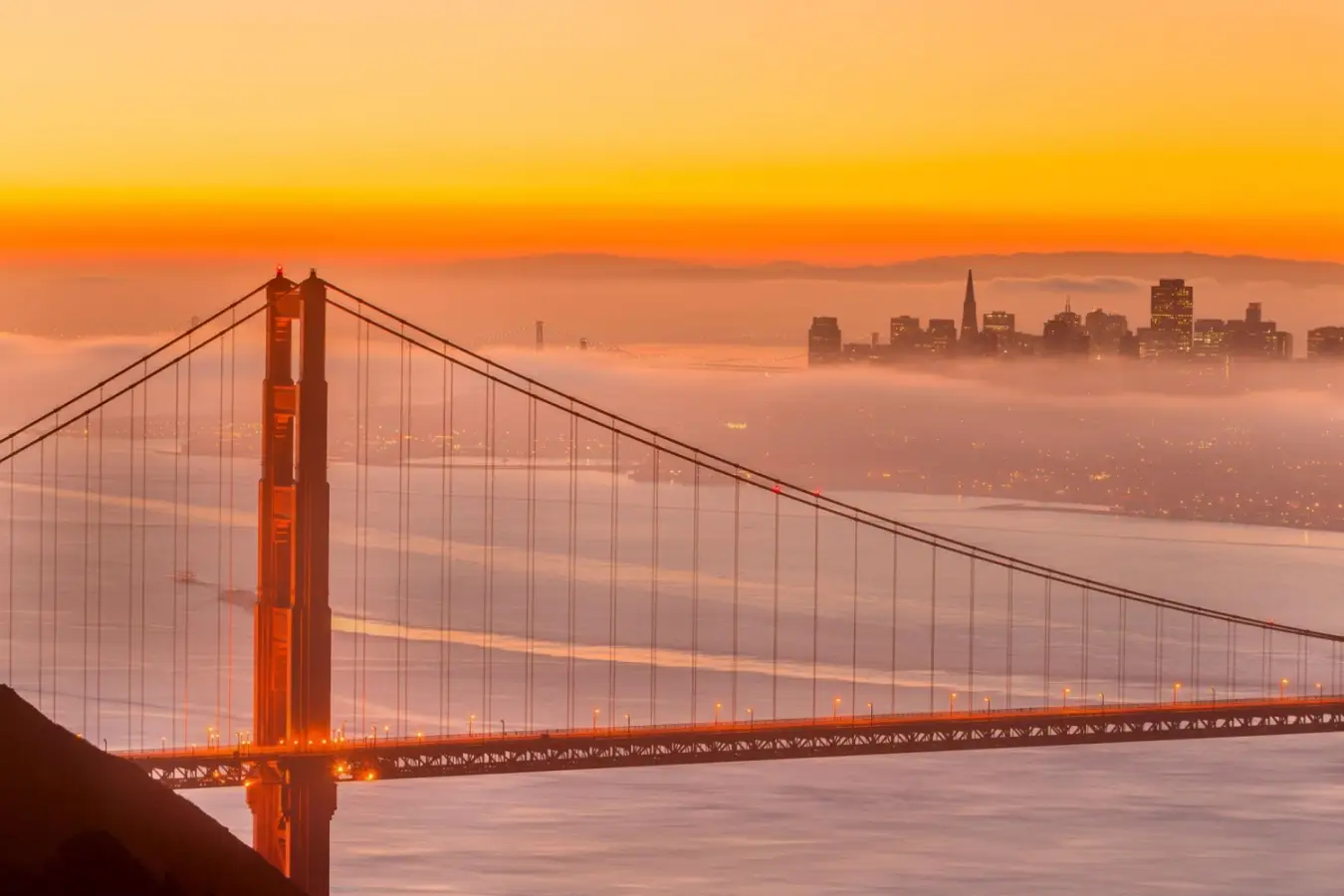 A panoramic photograph of the Golden Gate Bridge during sunrise, featuring thick fog covering the San Francisco skyline in the background.