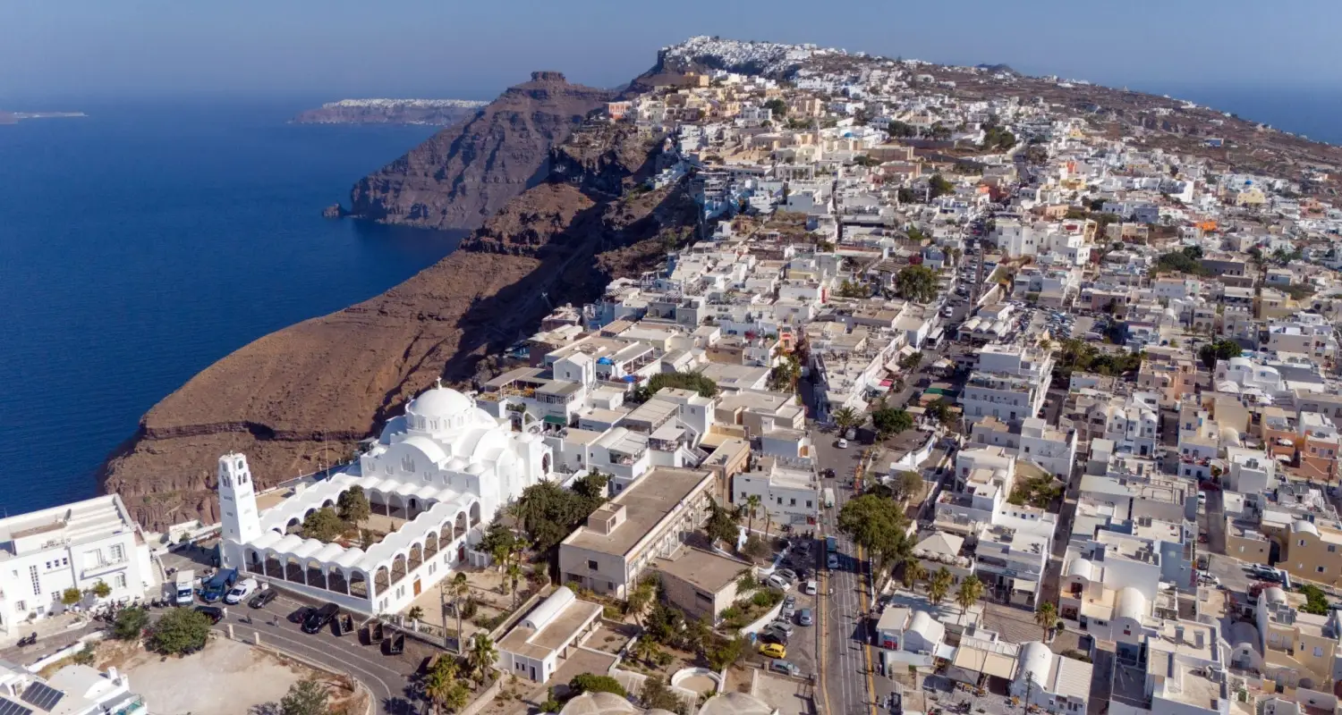 Aerial view of Fira, the cliffside capital of Santorini, Greece, featuring white Cycladic buildings, the Orthodox Metropolitan Cathedral, and the Aegean Sea caldera.