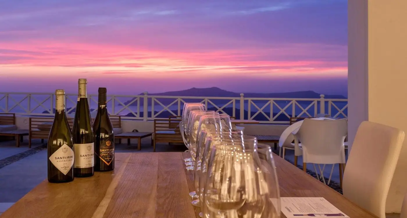 Wine bottles and rows of crystal glasses on an outdoor terrace overlooking the Santorini caldera during a purple sunset.