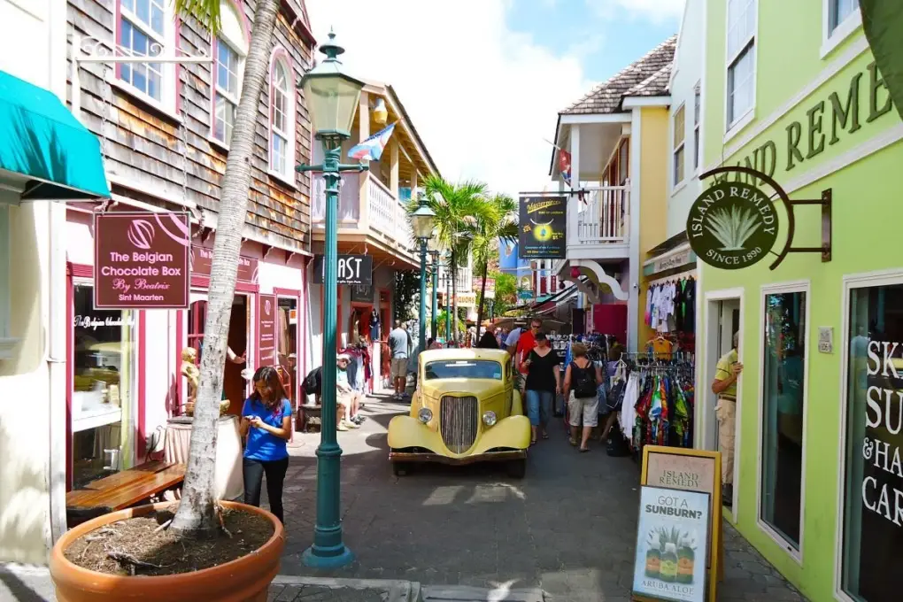 Pedestrian alleyway on Old Street in Philipsburg, Sint Maarten, with colorful storefronts, local duty-free shops, a vintage yellow car, and tourists walking under a sunny Caribbean sky.