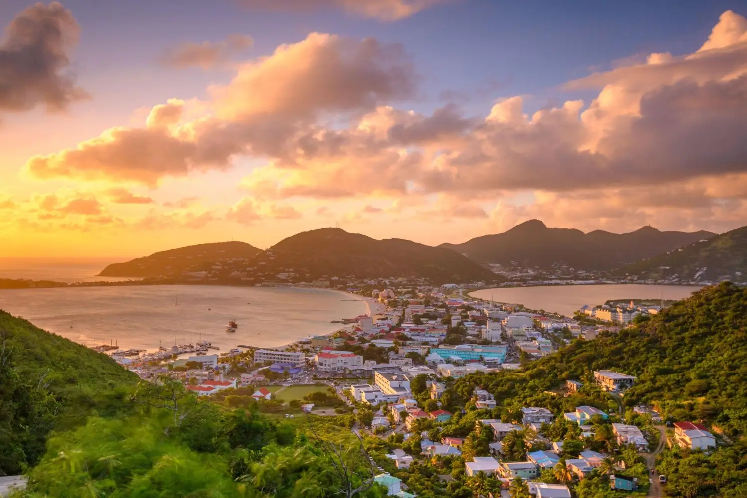 An aerial view of Philipsburg waterfront during a tropical sunset, showing a coastal town with a wide sandy beach, calm blue water, and rolling green mountains in the background.