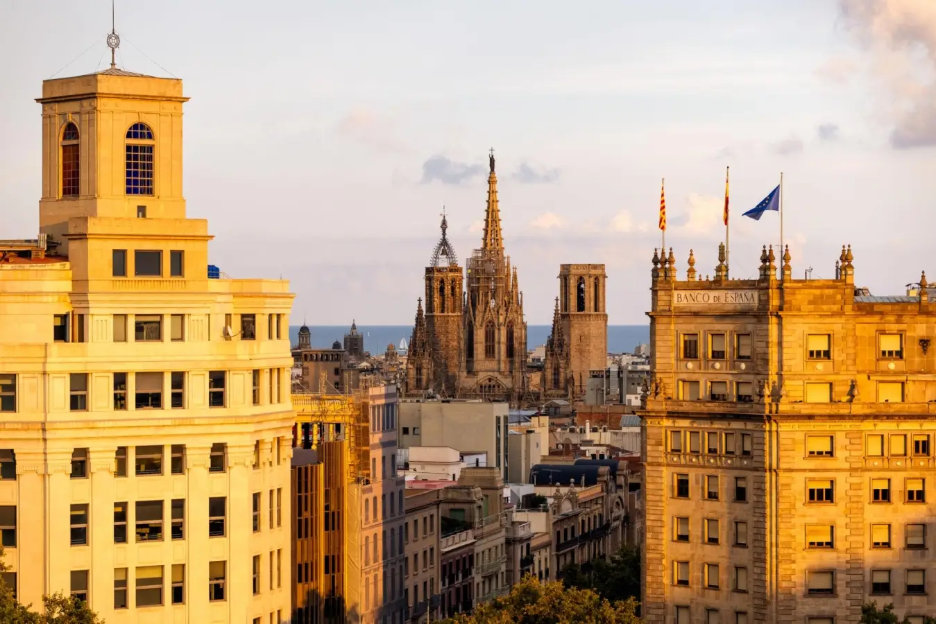 Rooftop view of the Barcelona skyline in Spain at golden hour, featuring the spires of the Barcelona Cathedral, the Banco de Espana building with flags, and historic architecture.