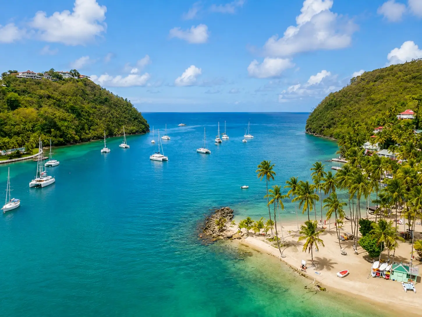 Aerial view of sailboats anchored in the turquoise waters of Marigot Bay, St. Lucia, surrounded by lush green hills and a sandy beach lined with palm trees.