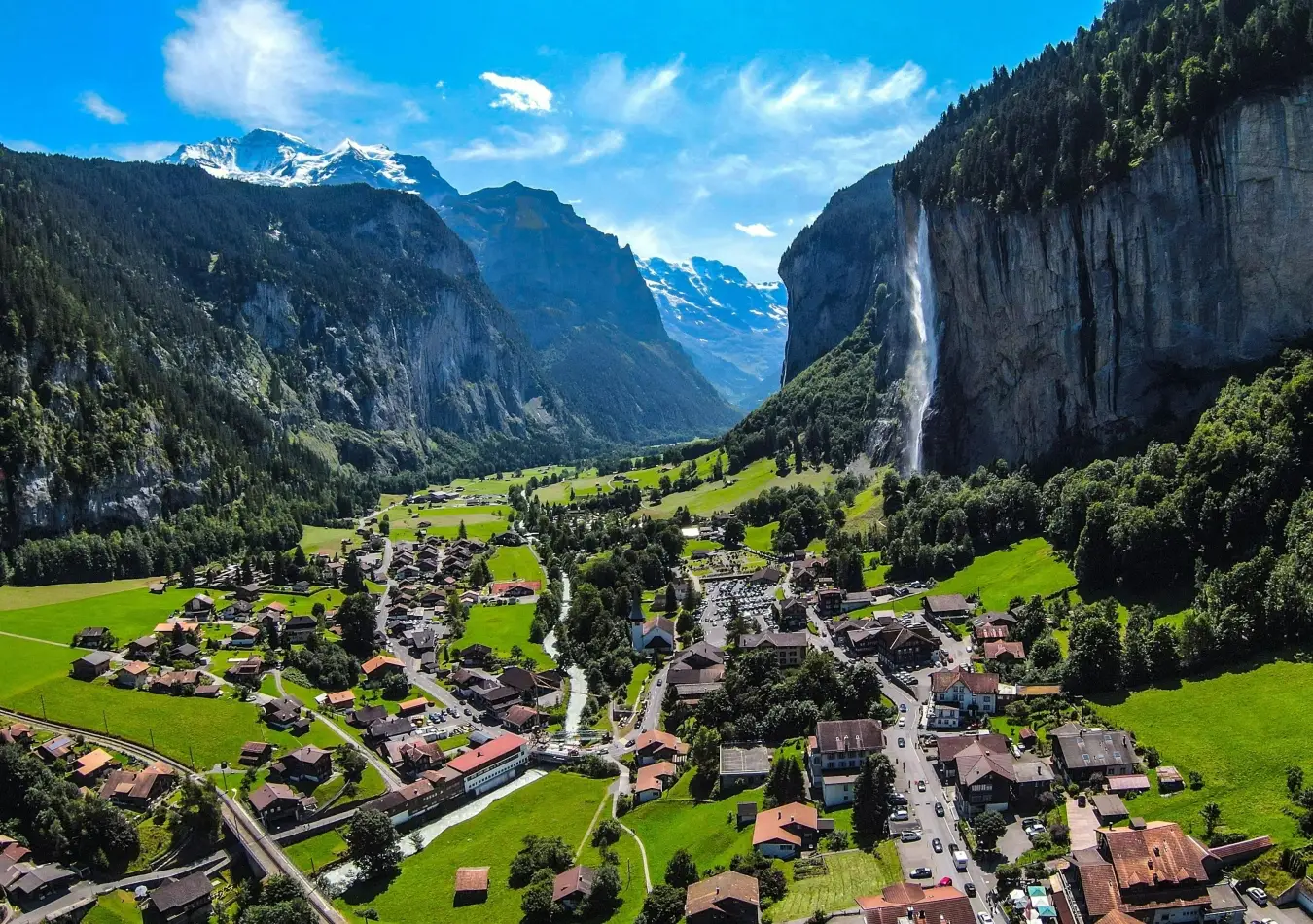 Aerial view of Lauterbrunnen village in Switzerland featuring Staubbach Falls crashing down steep rocky cliffs into a lush green alpine valley.