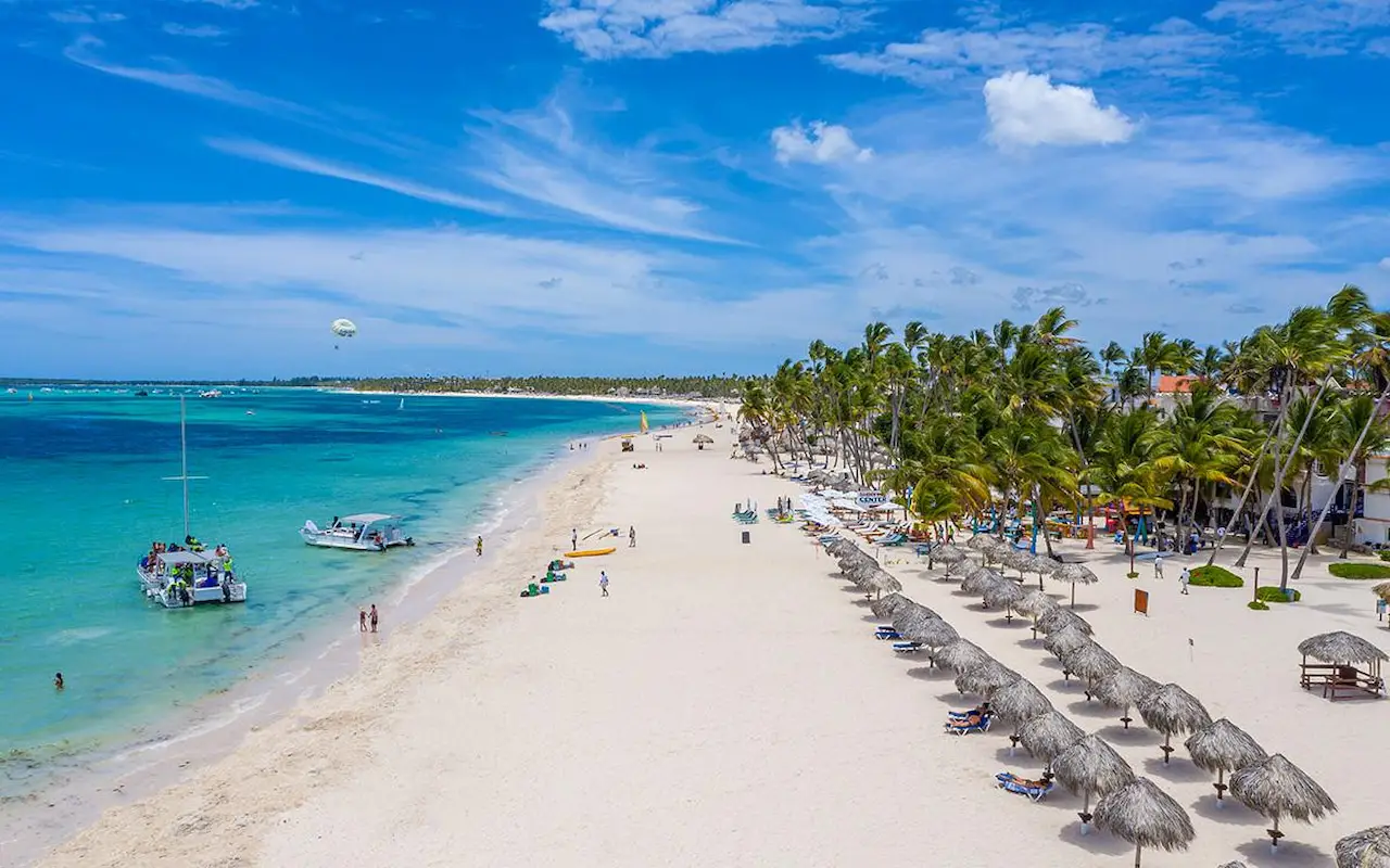 Aerial view of Bavaro Beach in Punta Cana with white sand, palm trees, thatched umbrellas, and boats in turquoise ocean water.