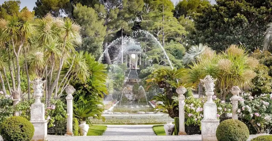 A large water fountain spraying in the center of the lush French Garden at Villa Ephrussi de Rothschild, framed by palm trees, white stone pillars and manicured hedges under a green forest canopy.